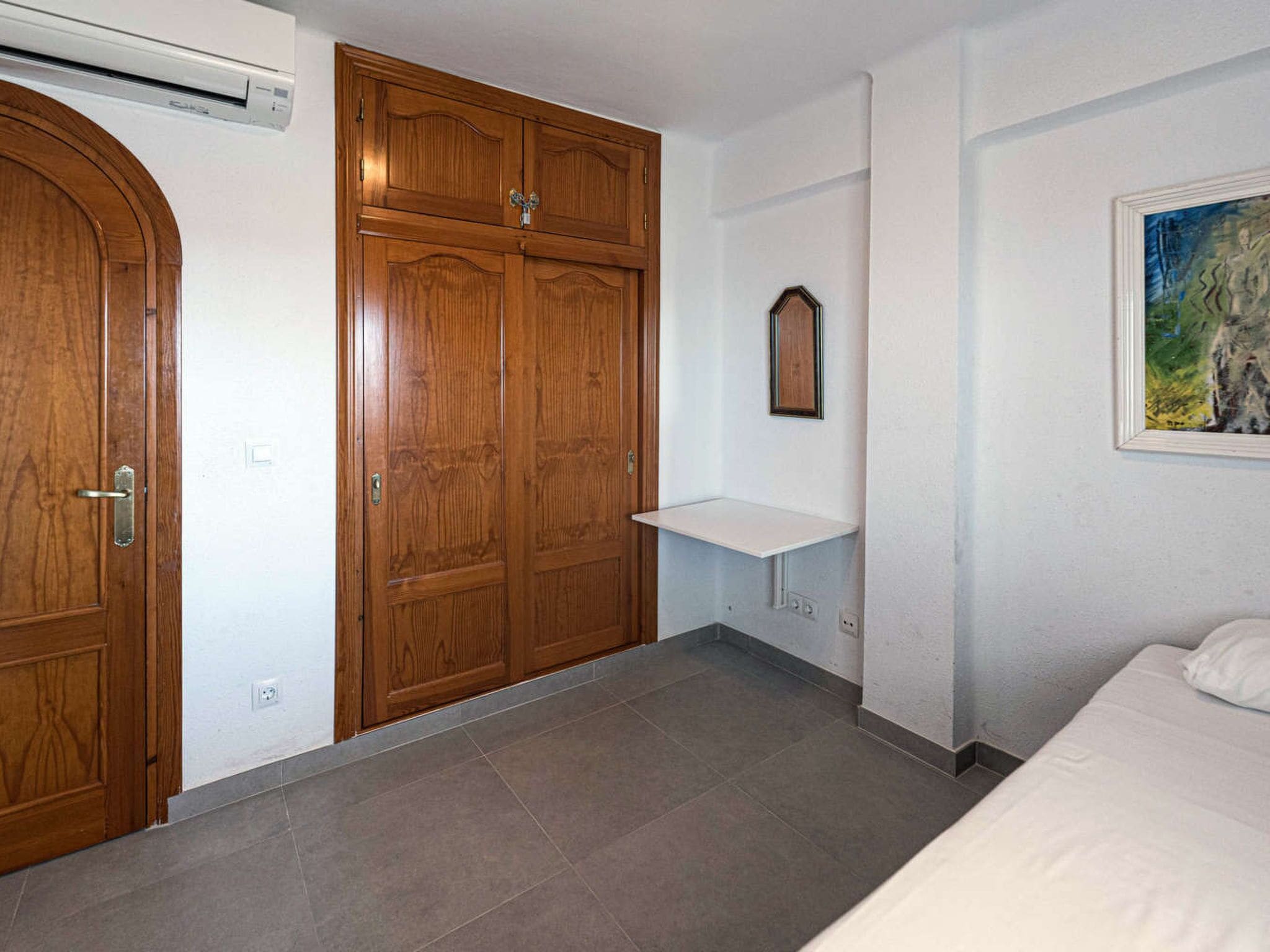 Quarto com cama, guarda-roupa de madeira, prateleira pequena e um quadro na parede.