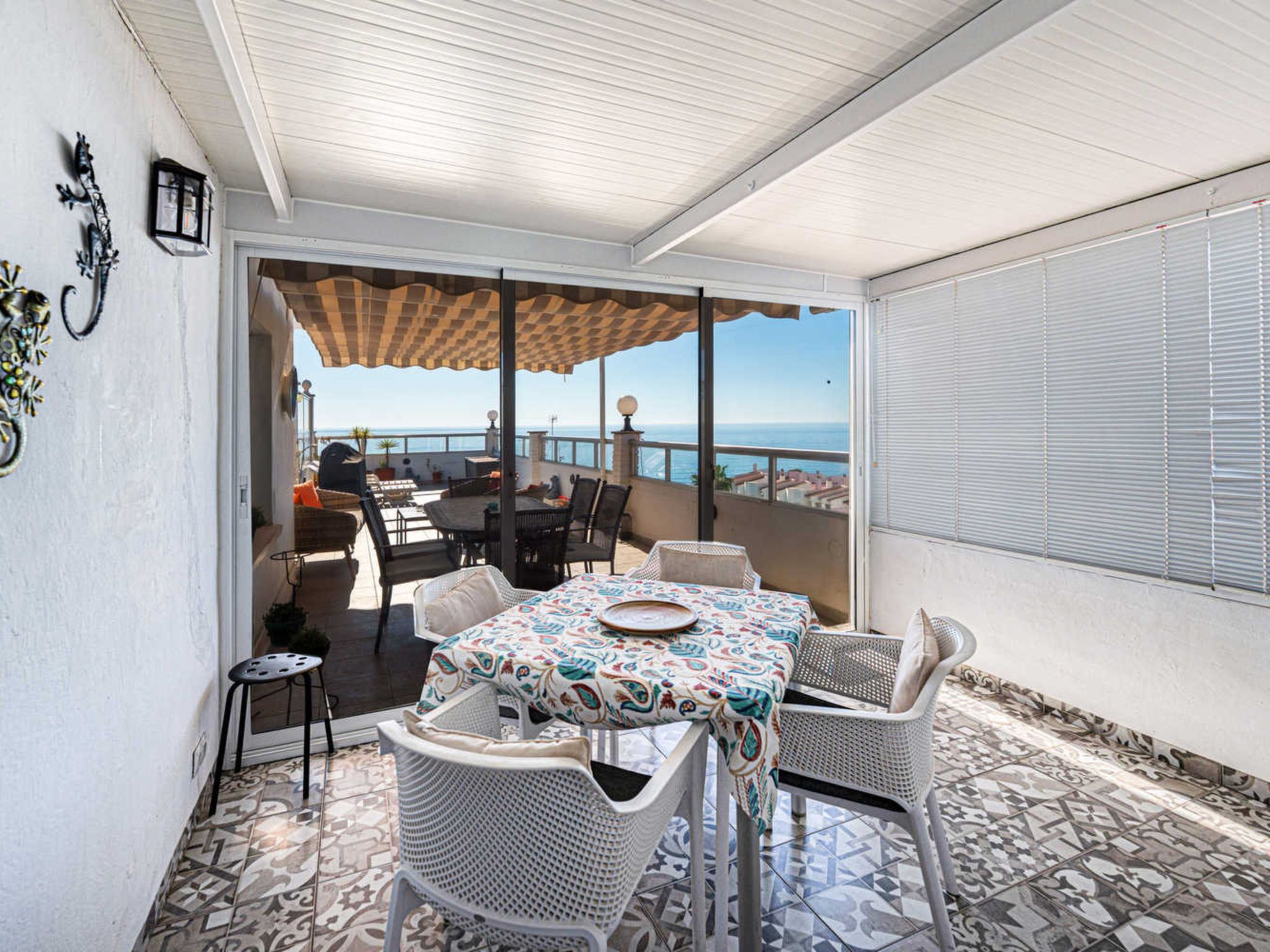 A covered outdoor seating area with a table and chairs, a view of the sea, and a tiled floor.