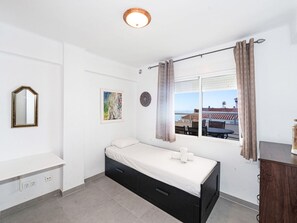 Um quarto pequeno e arrumado, com uma cama de solteiro, mesa de cabeceira, espelho e janela com cortinas.