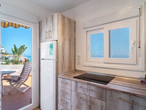 Cocina compacta con refrigerador empotrado, muebles de madera, cocina por inducción negra y ventana con vista al mar.