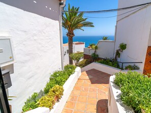 Un estrecho pasillo exterior con suelo de baldosas, rodeado de paredes blancas y vegetación, que ofrece vistas al mar.