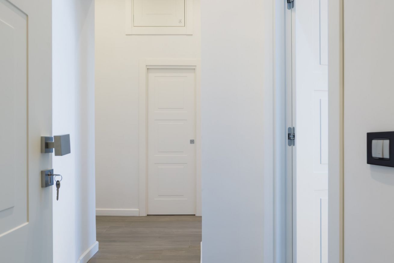 A hallway with a white door at the end, a light switch on the wall, and a key in the door lock.