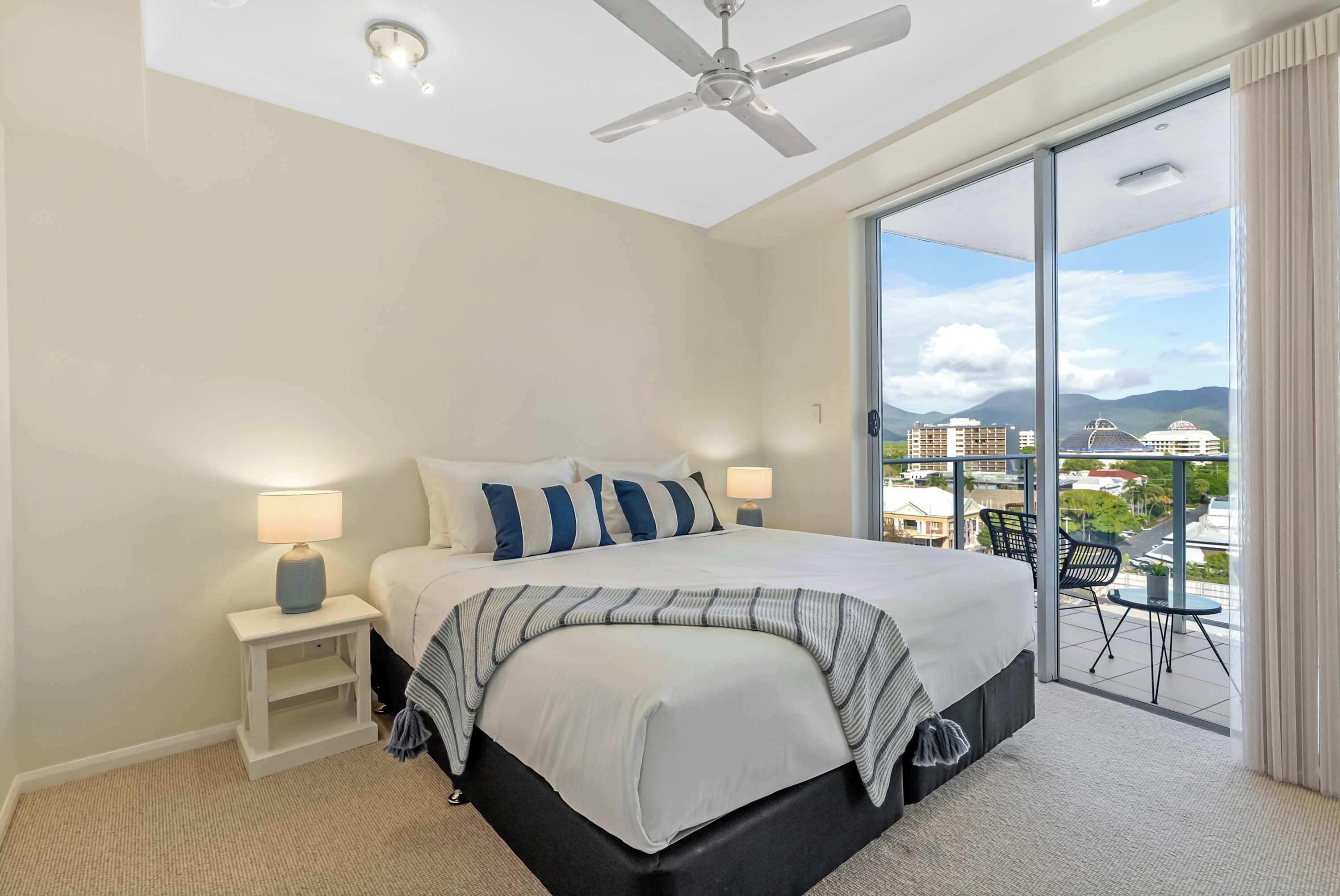 A bedroom with a bed, nightstands, a ceiling fan, and a view of the city through a large sliding glass door.
