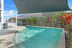 A swimming pool with a curved edge, surrounded by a tiled deck and a shaded area with a modern building in the background. - Seaview Escape—Scenic Views, Pool and Prime Location (Cairns City)