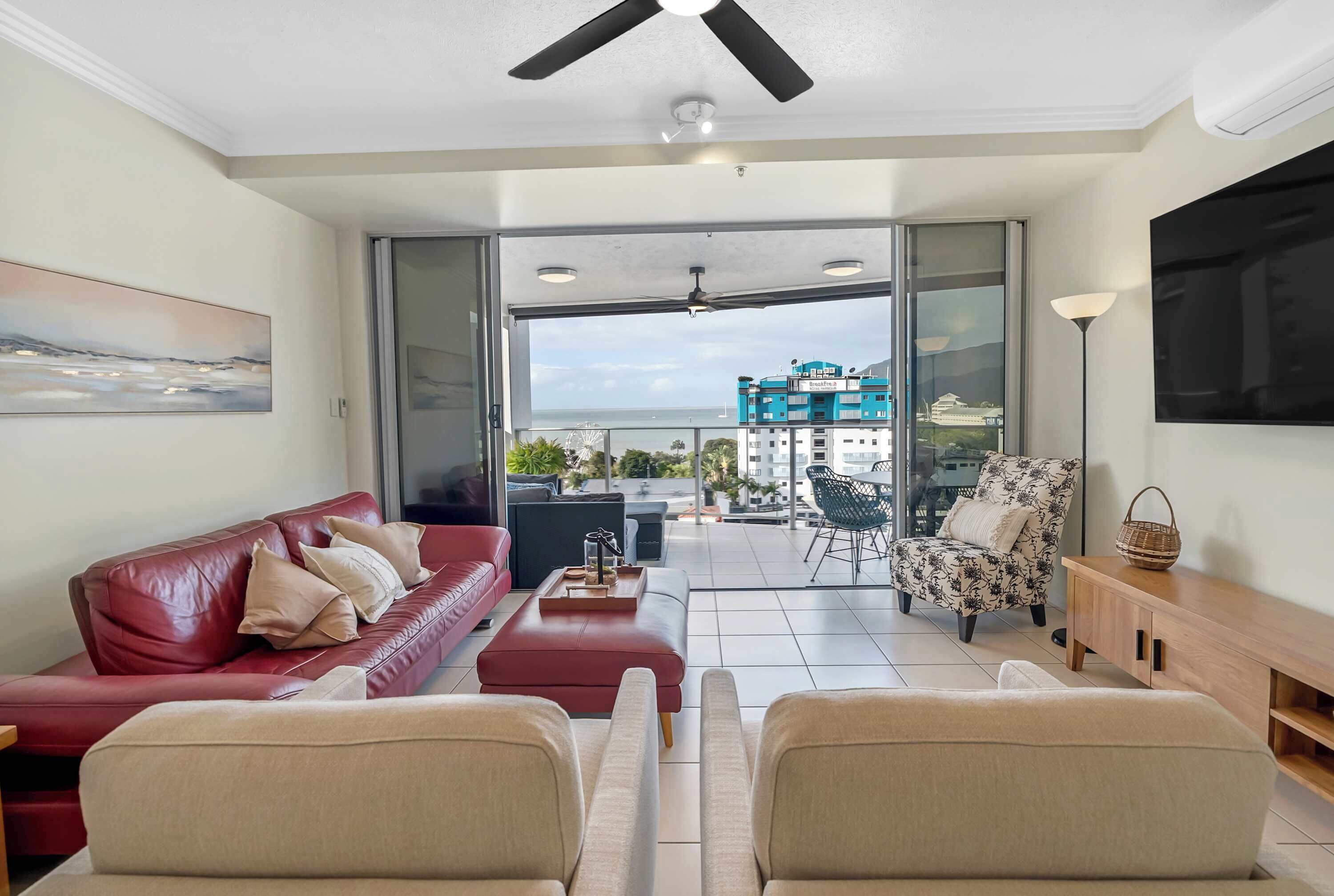 A modern living room with a red sofa, beige armchairs, a wooden side table, and a large window offering a view of a building and the sea.