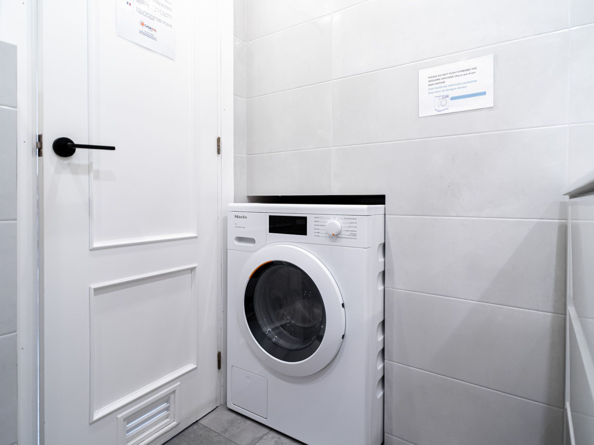 A compact, modern laundry room with a front-loading washing machine, a door with a handle, and a wall-mounted sign with instructions.