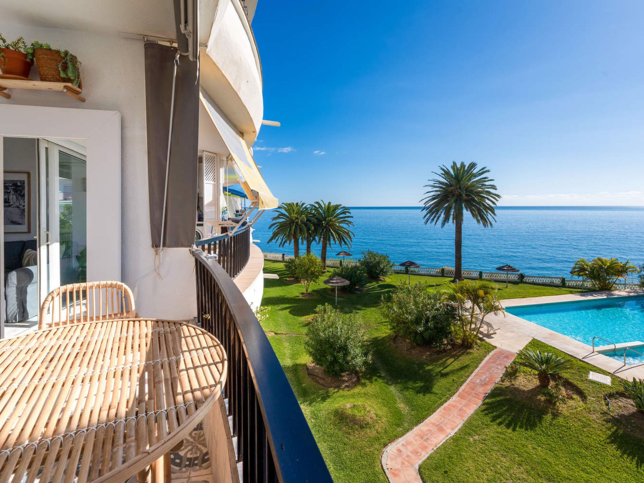 A balcony with a view of the sea, a pool, and a garden with palm trees.