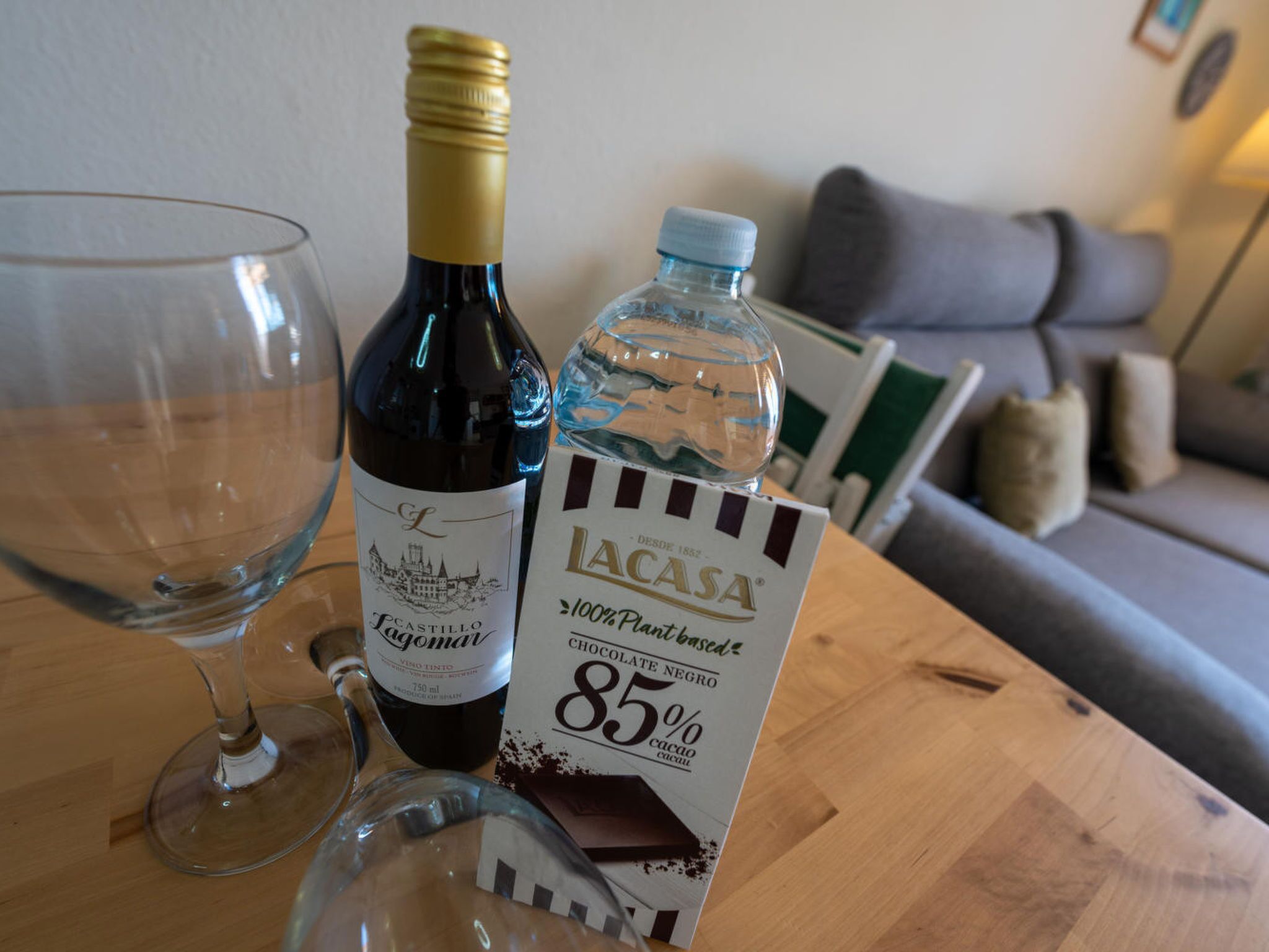 Une bouteille de vin, un verre et une bouteille d’eau sur une table en bois, avec un canapé en arrière-plan.