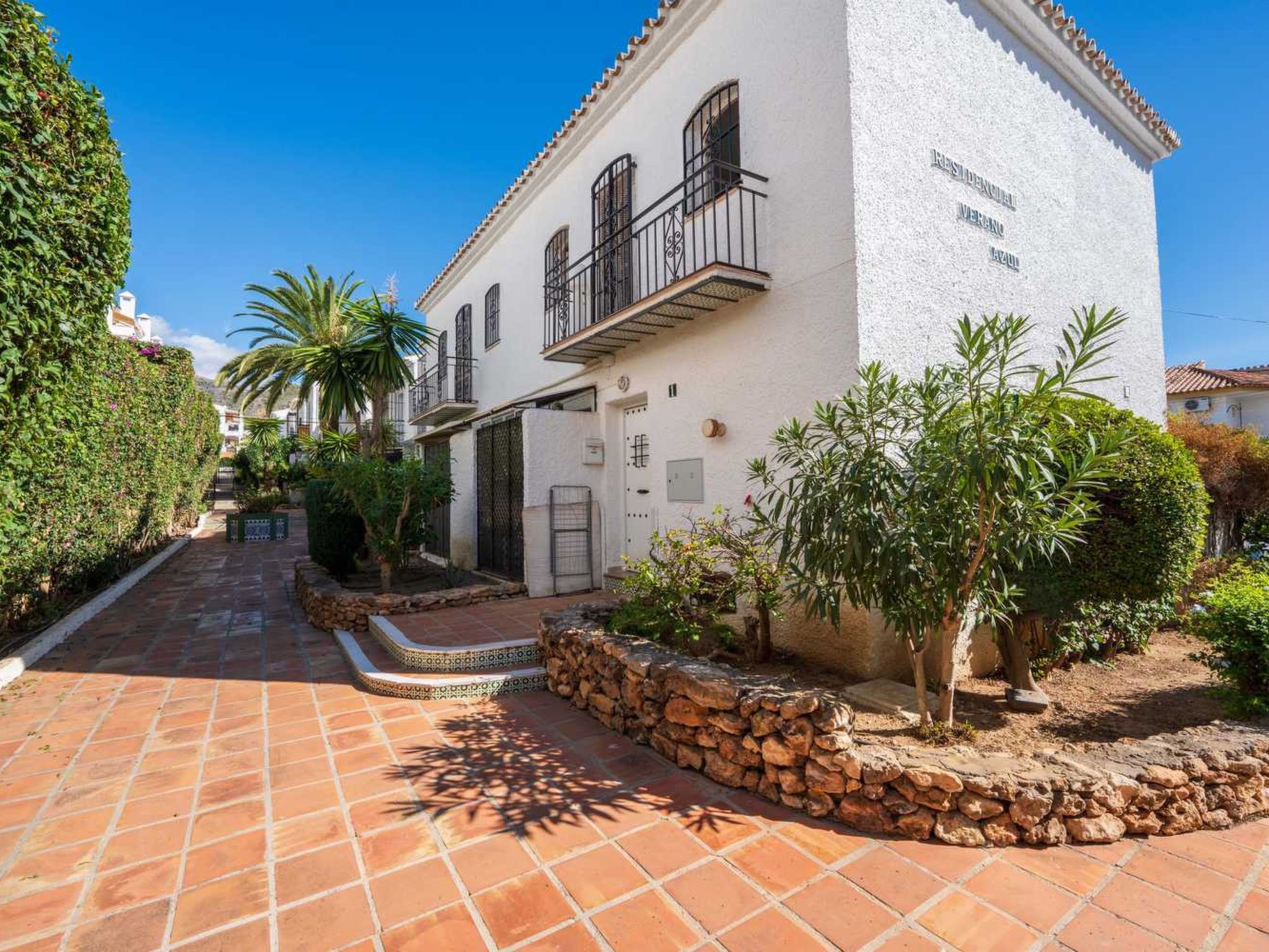 A white building with a balcony, a stone wall, and a tiled pathway.