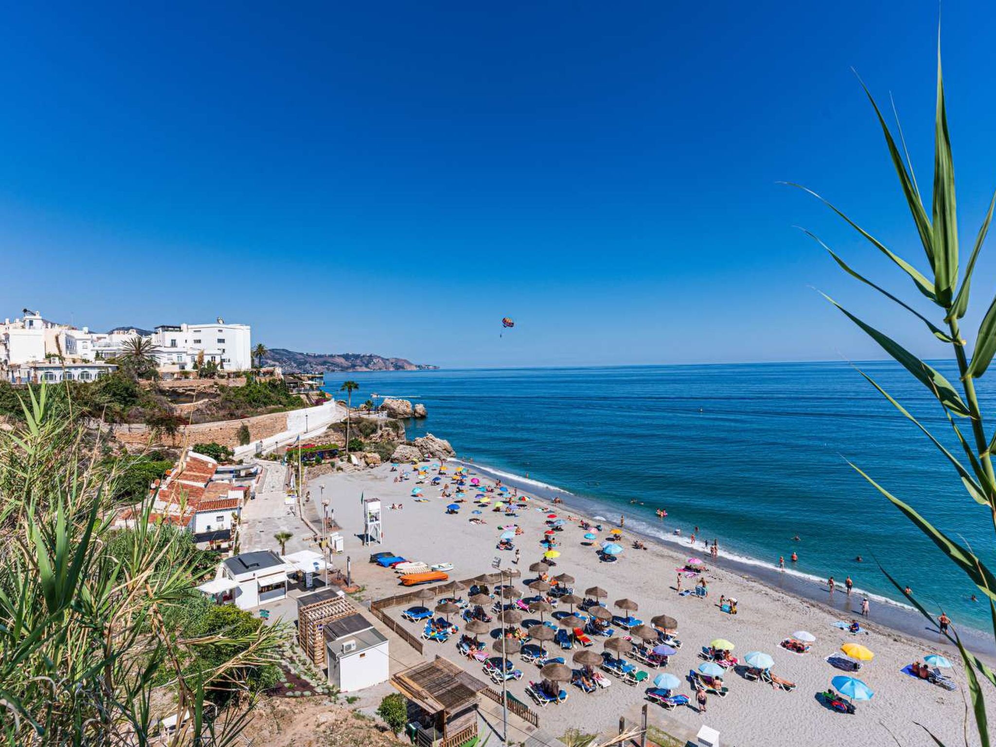 Une plage côtière avec un ciel dégagé, un parachute ascensionnel visible au loin, ainsi qu’une variété de parasols et de chaises longues.