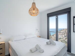 Ein Schlafzimmer mit Bett, Nachttisch, Lampe und Blick auf eine Küstenstadt.