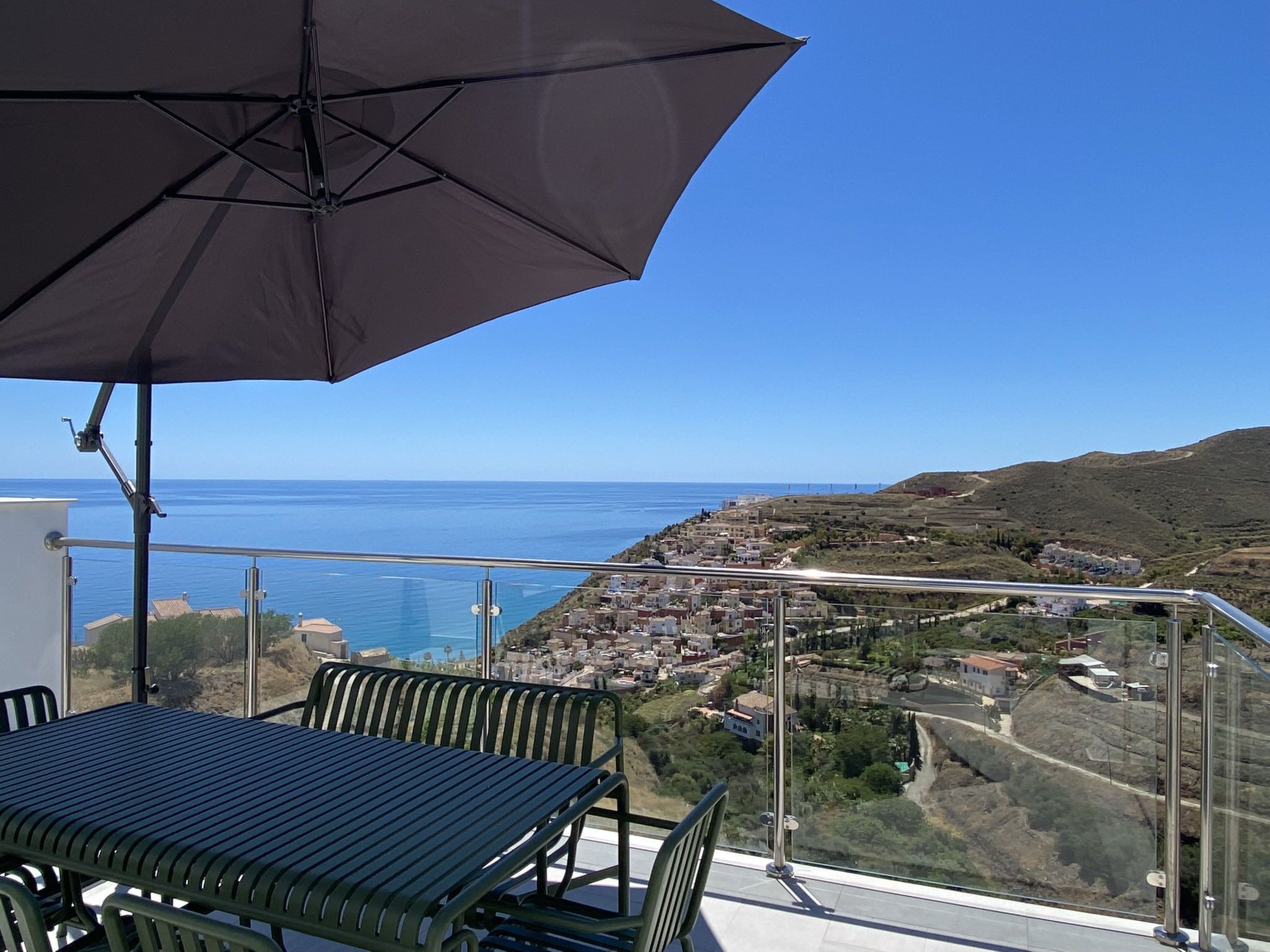 A balcony with a table and chairs, overlooking a coastal town and the sea.