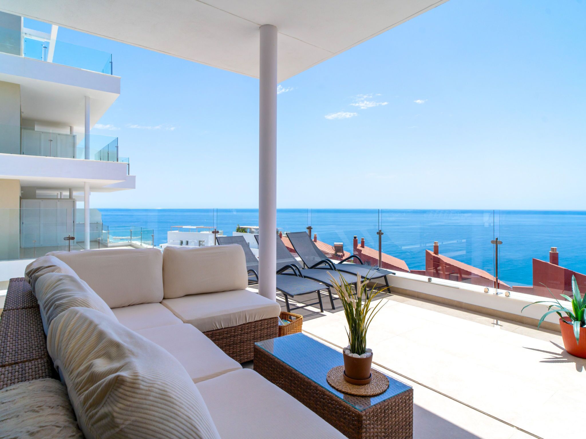 A modern balcony with a white sofa, lounge chairs, and a view of the sea.