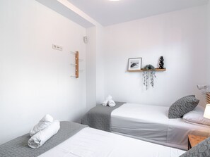 Habitación de hotel con dos camas, un estante en la pared con una planta y un cuadro, y una mesita de noche con una lámpara.