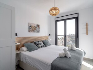 Une chambre moderne avec un lit, un luminaire suspendu, une fenêtre donnant sur la ville et un porte-serviettes fixé au mur.