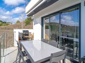 Ein moderner Essbereich im Freien mit einem weißen Tisch und Stühlen, einem Grill und einer Glasveranda mit Blick auf eine reizvolle Hügellandschaft.