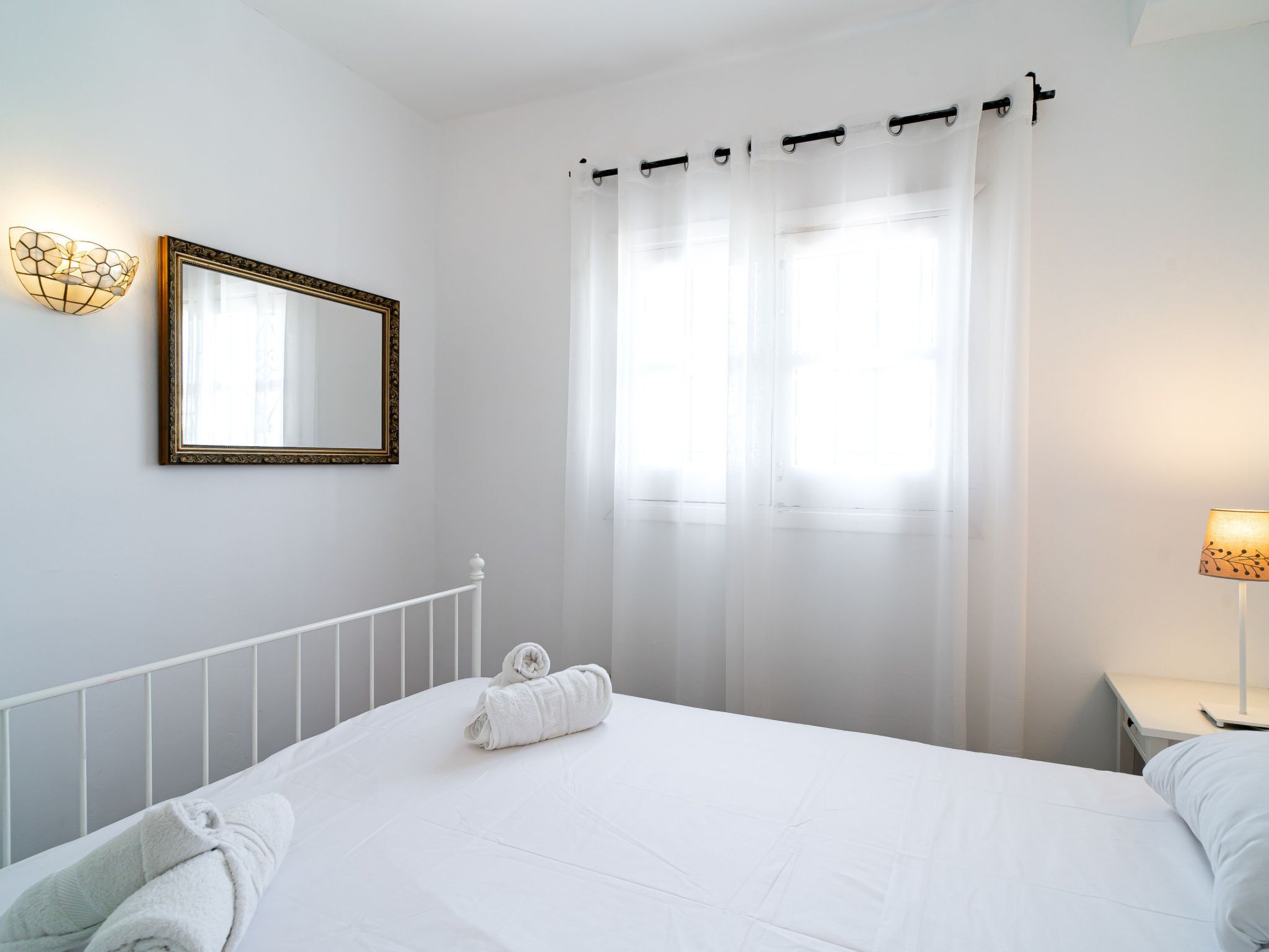 A hotel room with a bed, a wall-mounted mirror, a window with sheer curtains, and a bedside lamp.