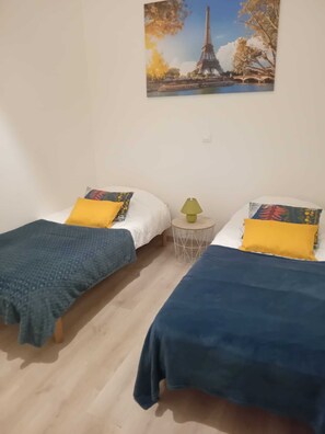 Ein Zimmer mit zwei Betten, jeweils mit einer blauen Bettdecke und einem gelben Kissen. Zwischen den Betten steht ein kleiner runder Tisch mit einer Lampe. An der Wand hängt ein Bild vom Eiffelturm.