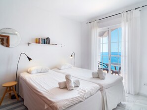 A hotel room with two beds, a small wooden stool, a bookshelf with books, a round mirror, and a view of the sea through a large window. - Villa Prime Seaview by Casasol by Interhome (Nerja)
