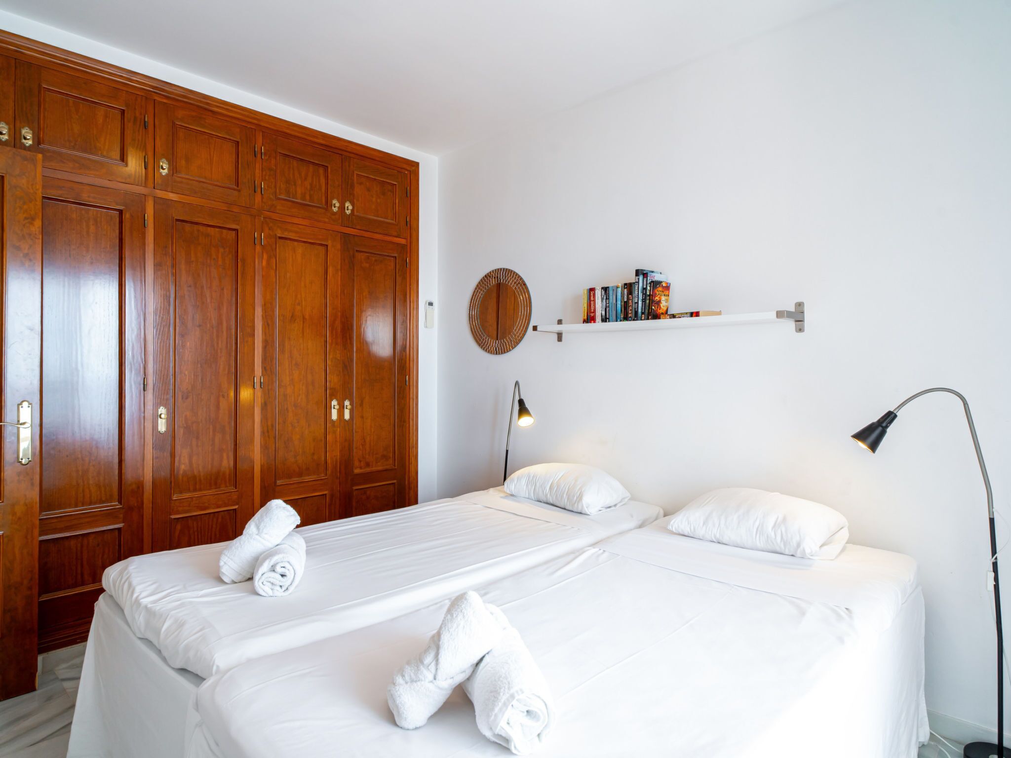 Hotelkamer met twee bedden, een houten garderobe, een ronde wanddecoratie, een plank met boeken en een bureaulamp.