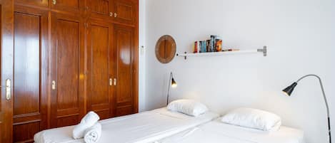 A hotel room with two beds, wooden wardrobe, a round wall decoration, a shelf with books, and a desk lamp.