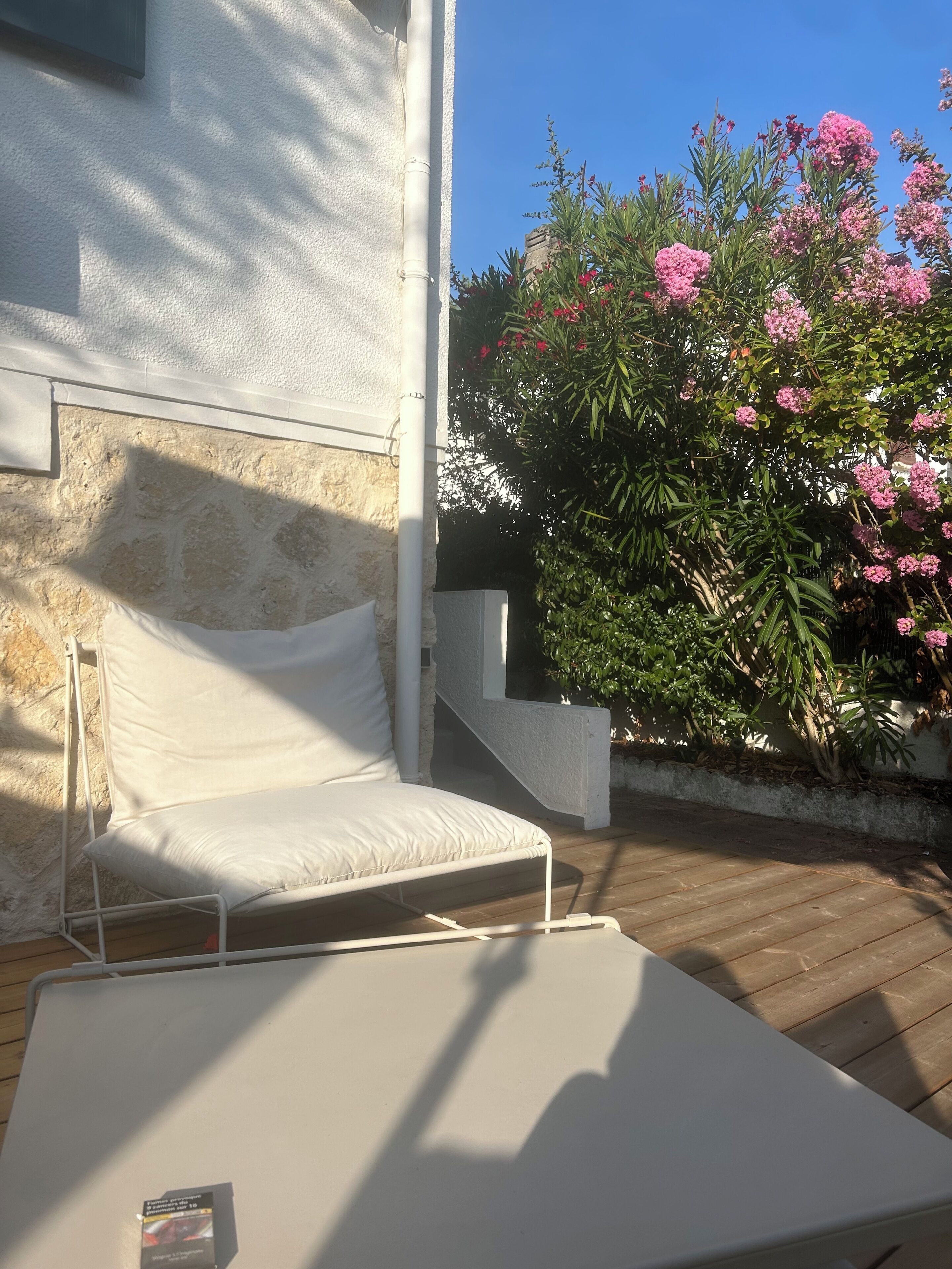 A sunlit outdoor seating area with a white cushioned bench, a wooden deck, and a stone wall.