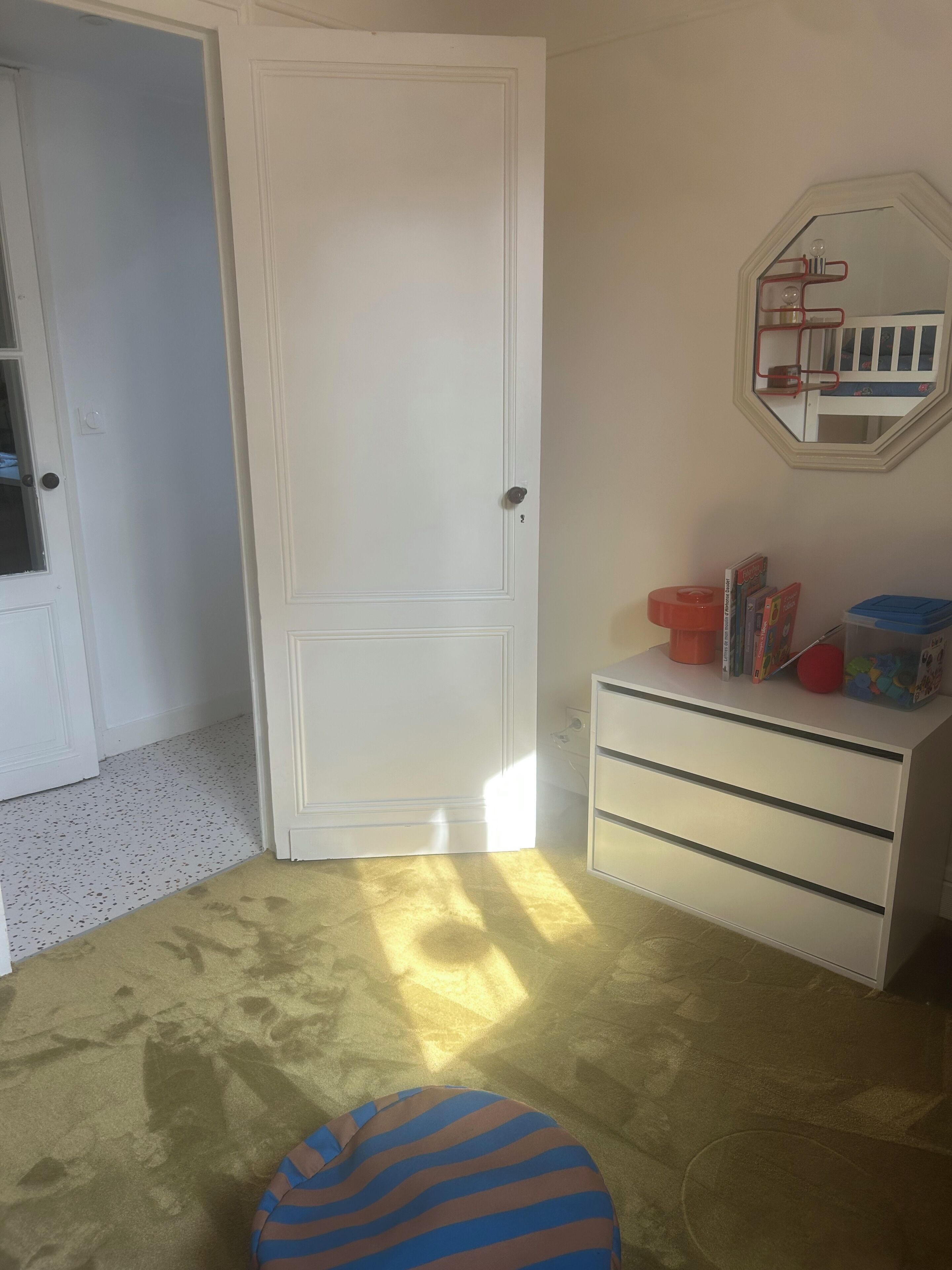 Een kamer met een gesloten witte deur, een witte dressoir met lades, een klein rond poef en een spiegel aan de muur.