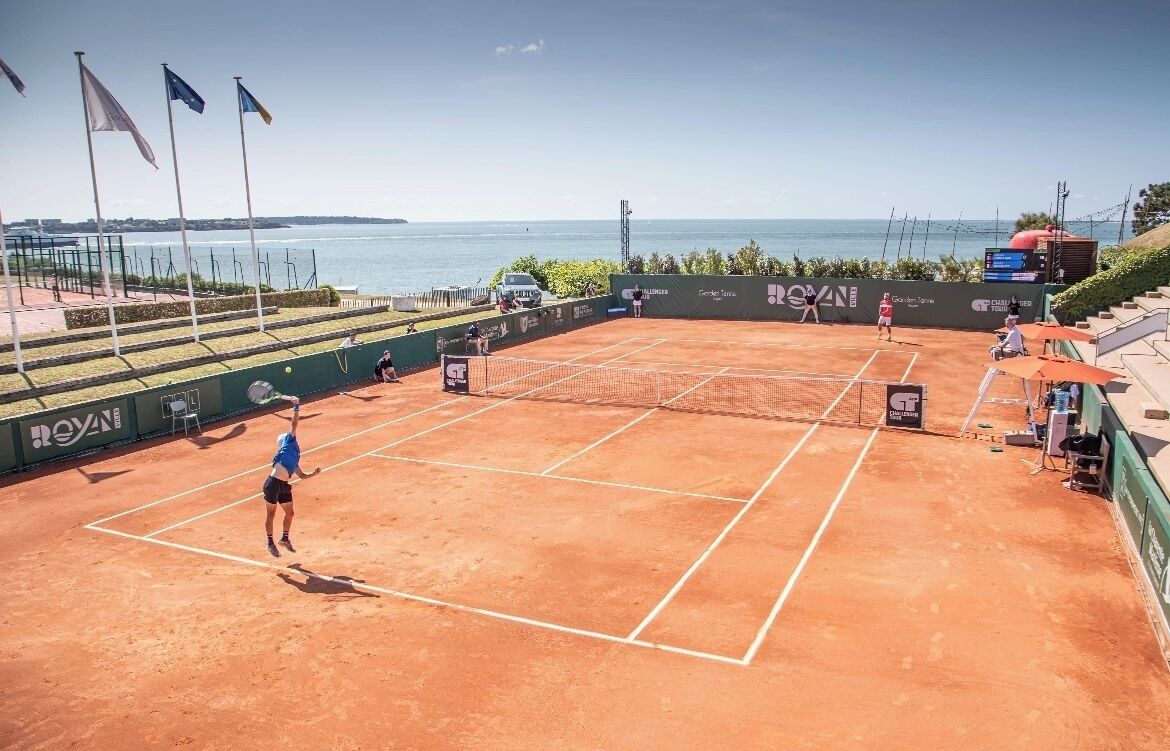 A clay tennis court with a net, surrounded by a green fence, and flags on poles.