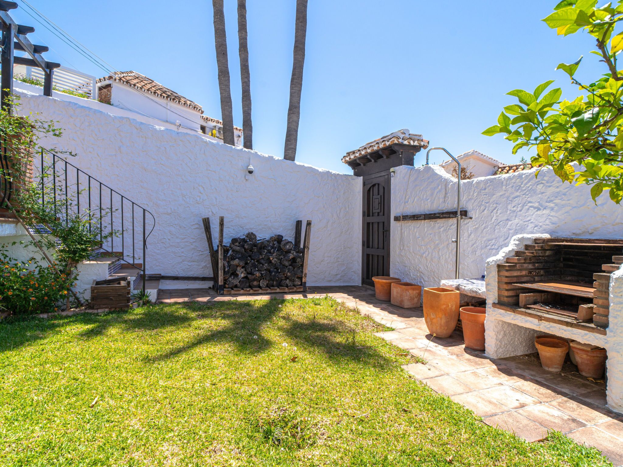 Un patio con un muro blanco, una escalera de madera, un portón de metal y macetas de terracota.