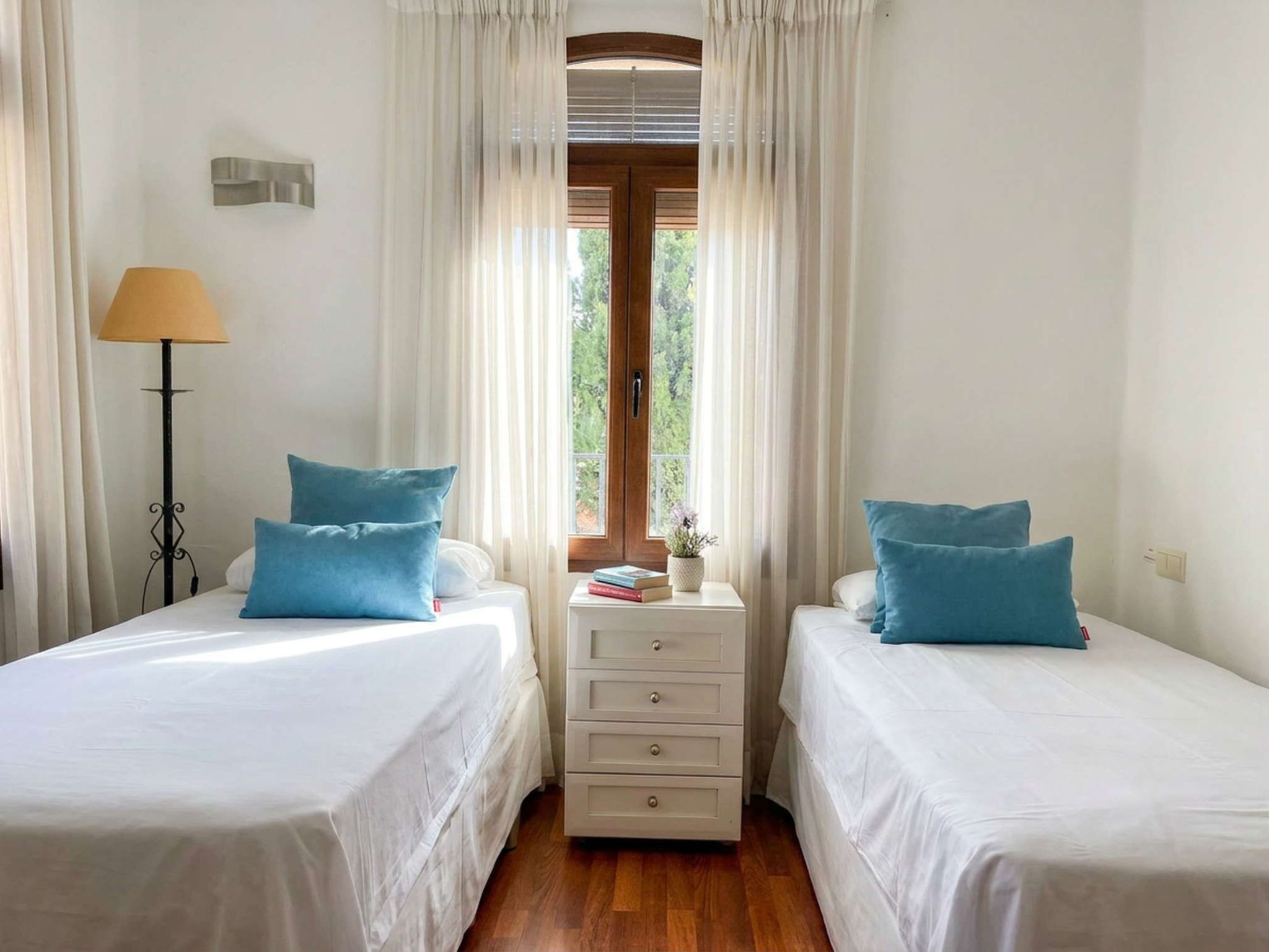 Ein Zimmer mit zwei Betten, einem Nachttisch mit Schubladen, einer Lampe und einem Fenster mit Vorhängen.
