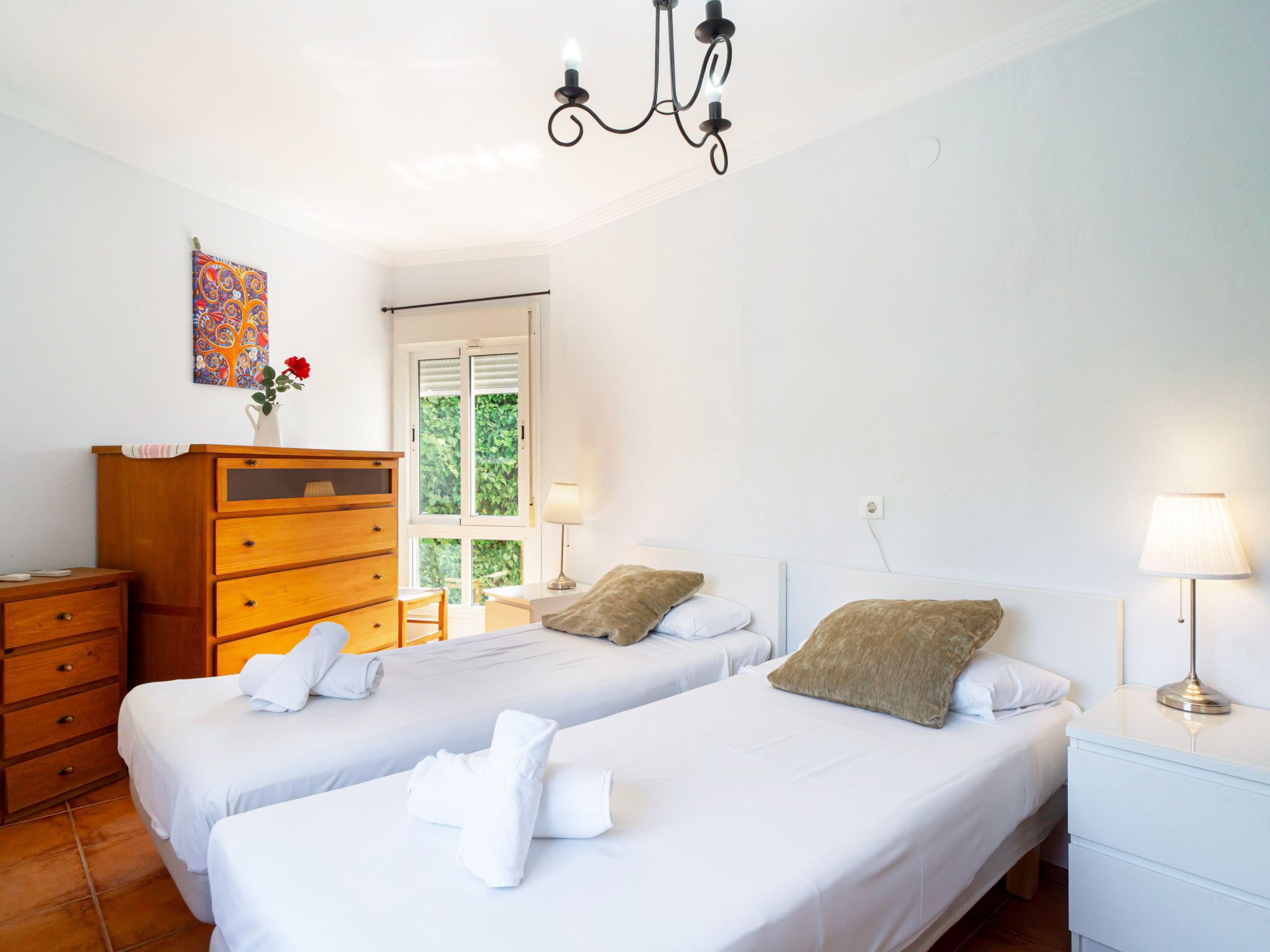 A hotel room with two beds, a wooden dresser, a window with a view of greenery, and a chandelier.