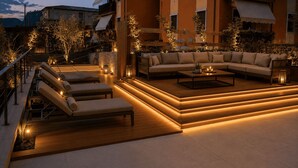 Un'area relax esterna moderna con divani, poltrone e un tavolino, circondata da piante e illuminazione soffusa.
