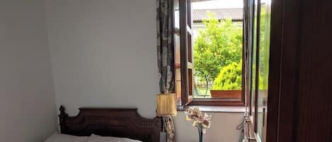 A bedroom with a wooden ceiling, a bed with a white duvet, a nightstand with a lamp, and a window with a view of greenery.