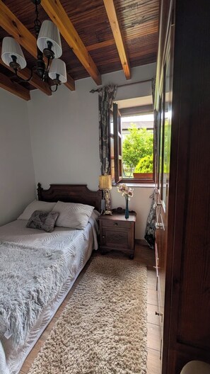 A bedroom with a wooden ceiling, a bed with a white duvet, a nightstand with a lamp, and a window with a view of greenery.