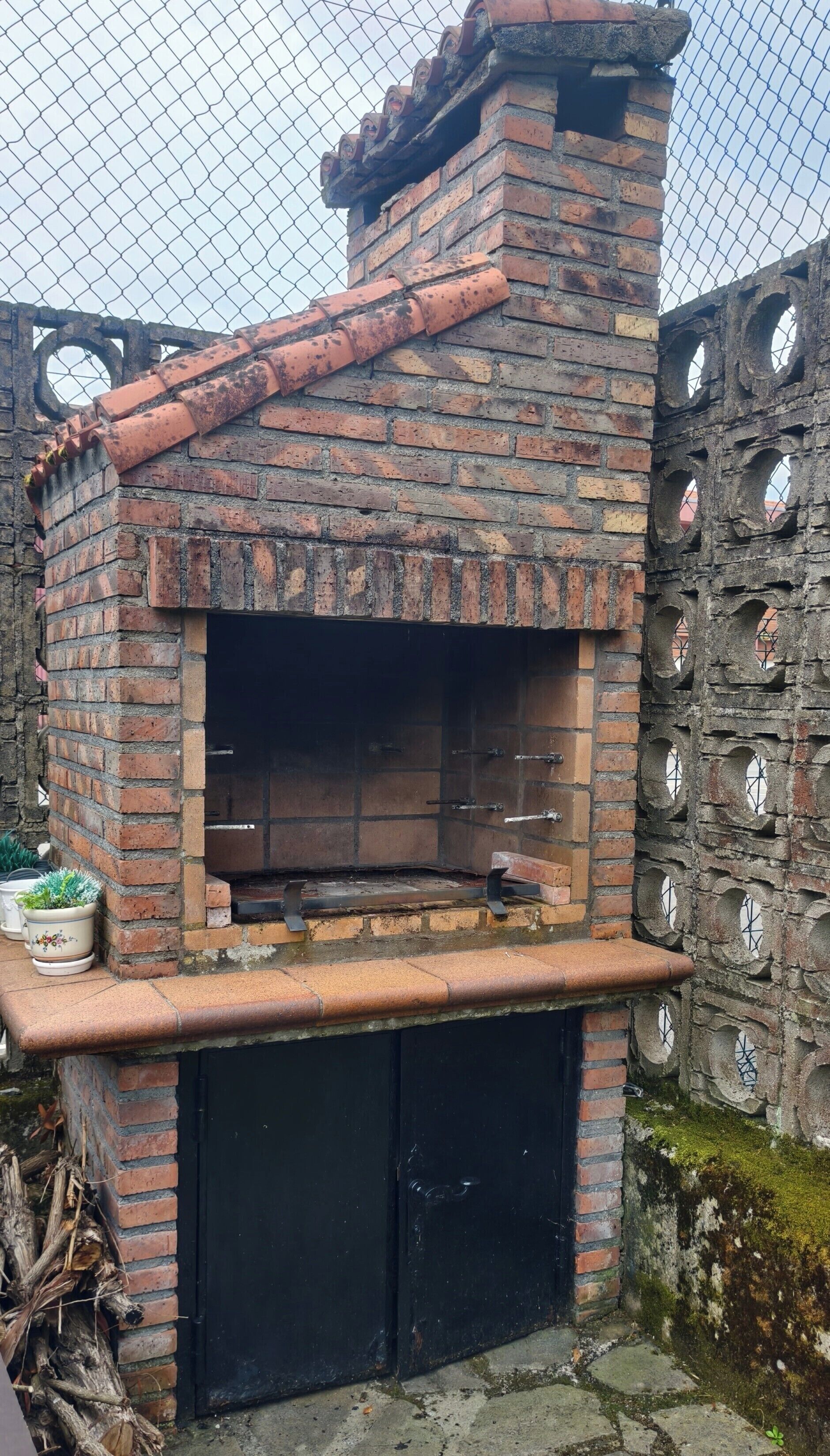 Un horno de ladrillo al aire libre con puerta negra, rodeado por una cerca de cadena y un muro de piedra con aberturas circulares.