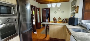 Uma cozinha com geladeira de aço inoxidável, micro-ondas e máquina de café. Há uma mesa de jantar de madeira com cadeiras e uma pequena ilha com pia.
