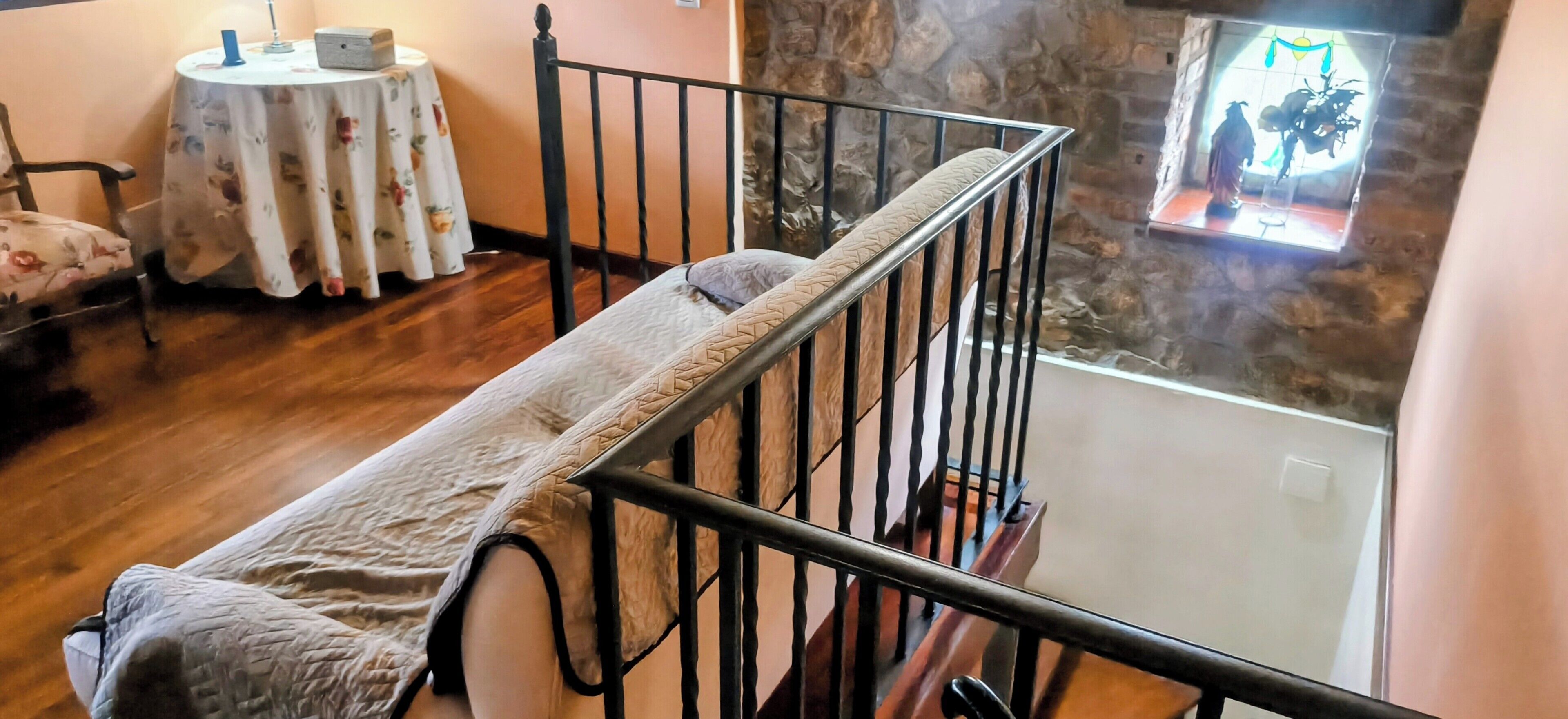 Una habitación con piso de madera, una cama con cabecera, una mesita redonda con mantel floral y una pared de piedra con un ventanal decorativo.