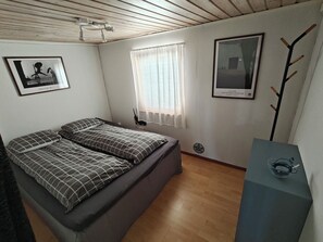 A room with a bed, a window with curtains, a wall-mounted light fixture, and two framed pictures.