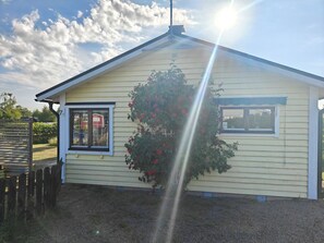 A single-story yellow house with a gabled roof, a wooden fence, and a bush with red flowers.