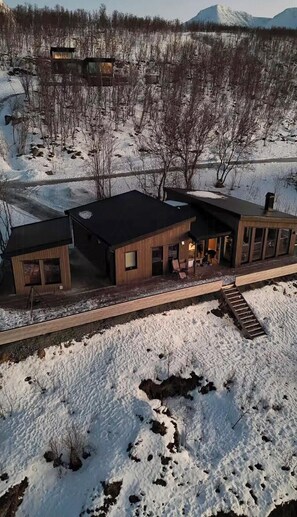 A modern cabin with a wooden exterior, a covered patio, and a view of a snowy landscape with mountains in the background.