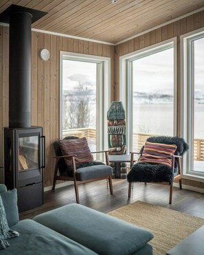 A cozy living room with a wood-burning stove, two chairs with patterned cushions, a sofa, and large windows offering a view of a body of water.
