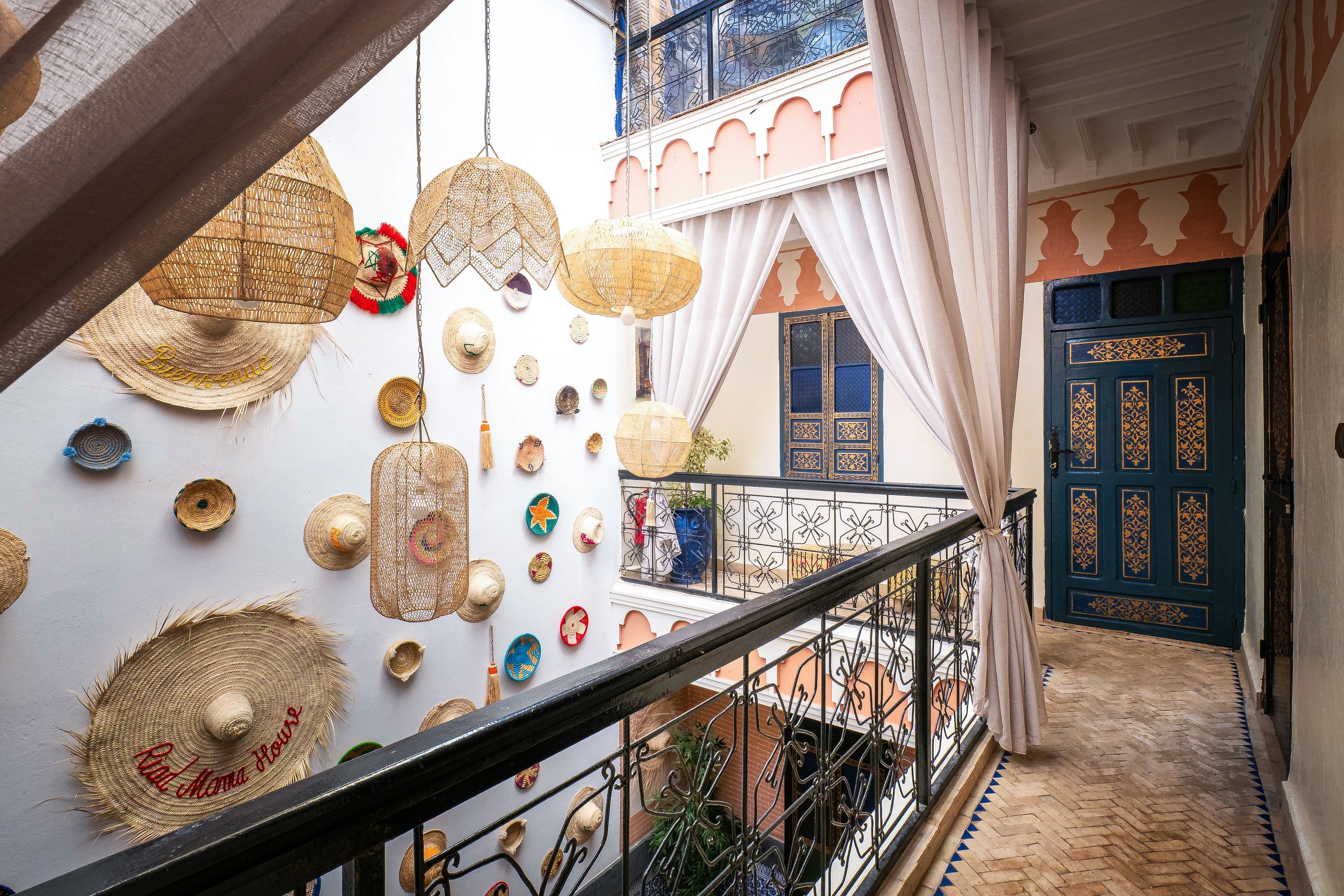 A balcony with a view of a blue door with intricate patterns, a white wall with various woven items, and a metal railing.