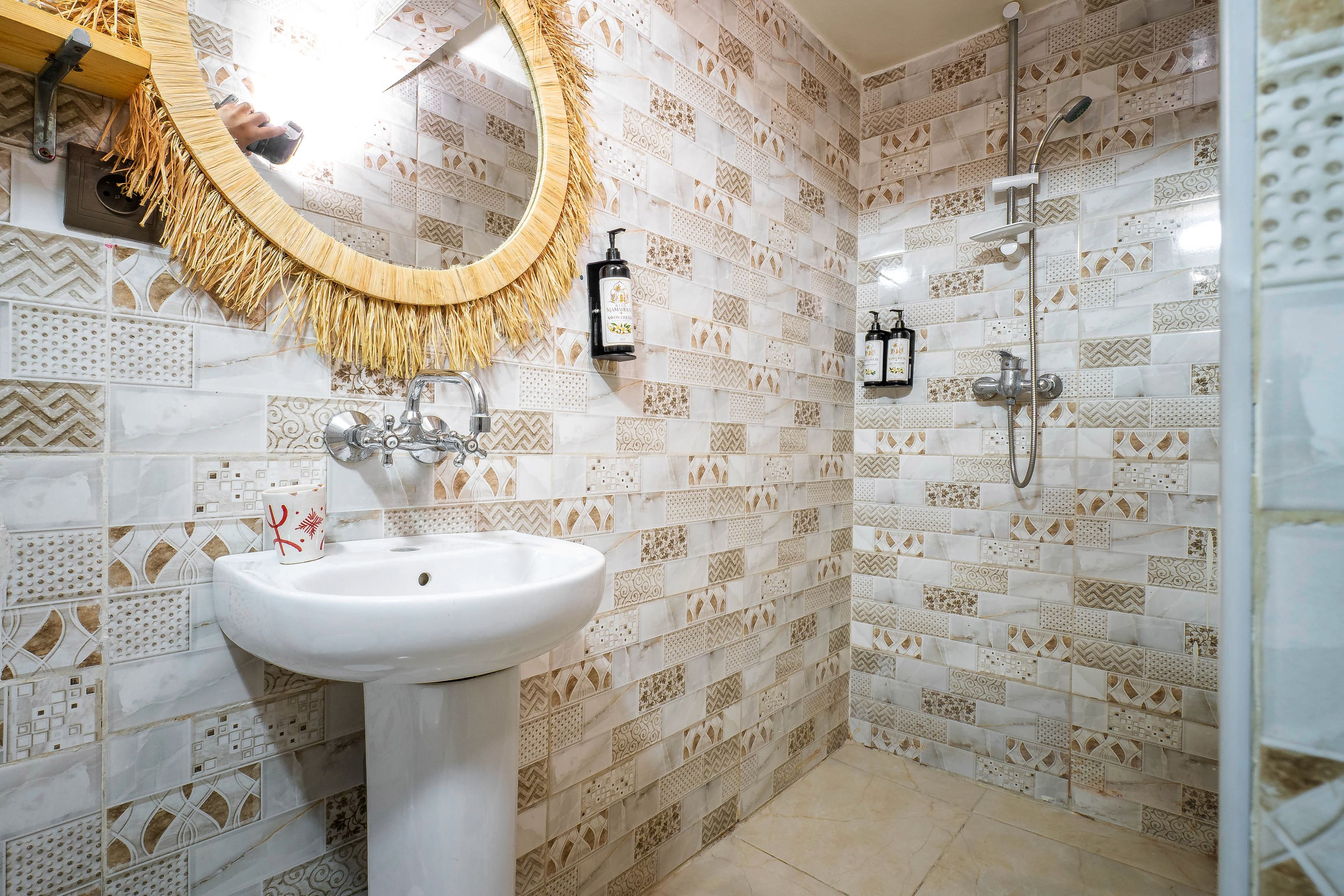 Une salle de bain avec un lavabo blanc, un miroir orné d’une frange en raphia et un espace douche avec des carreaux à motifs.