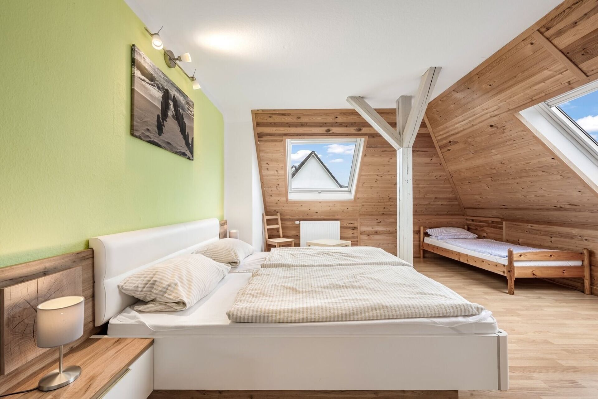 Une chambre avec un grand lit, un lit plus petit, un bureau et une chaise. Il y a une fenêtre donnant sur un immeuble et une lucarne.