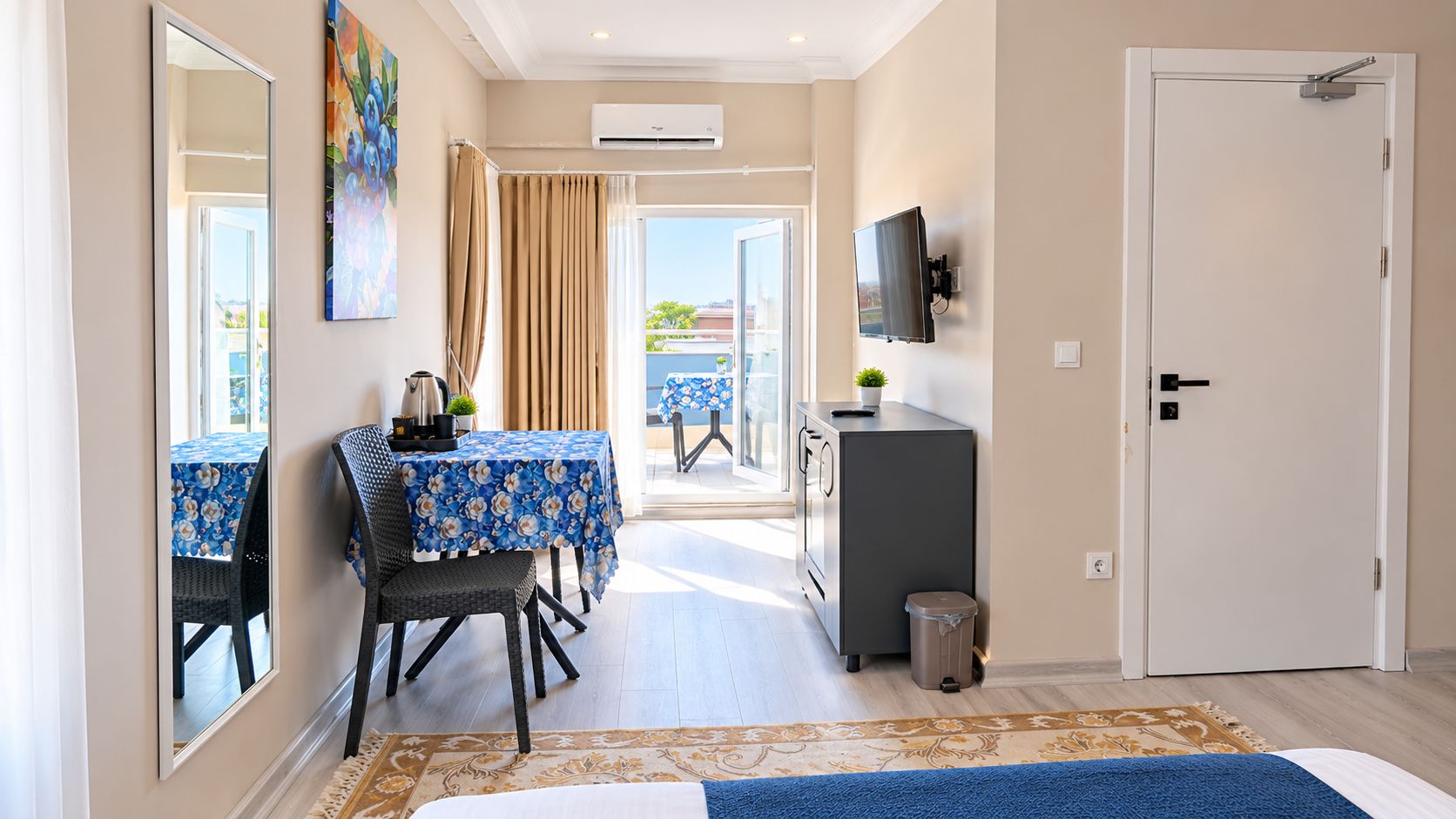 A hotel room with a dining area, a TV mounted on the wall, and a door leading to a balcony.