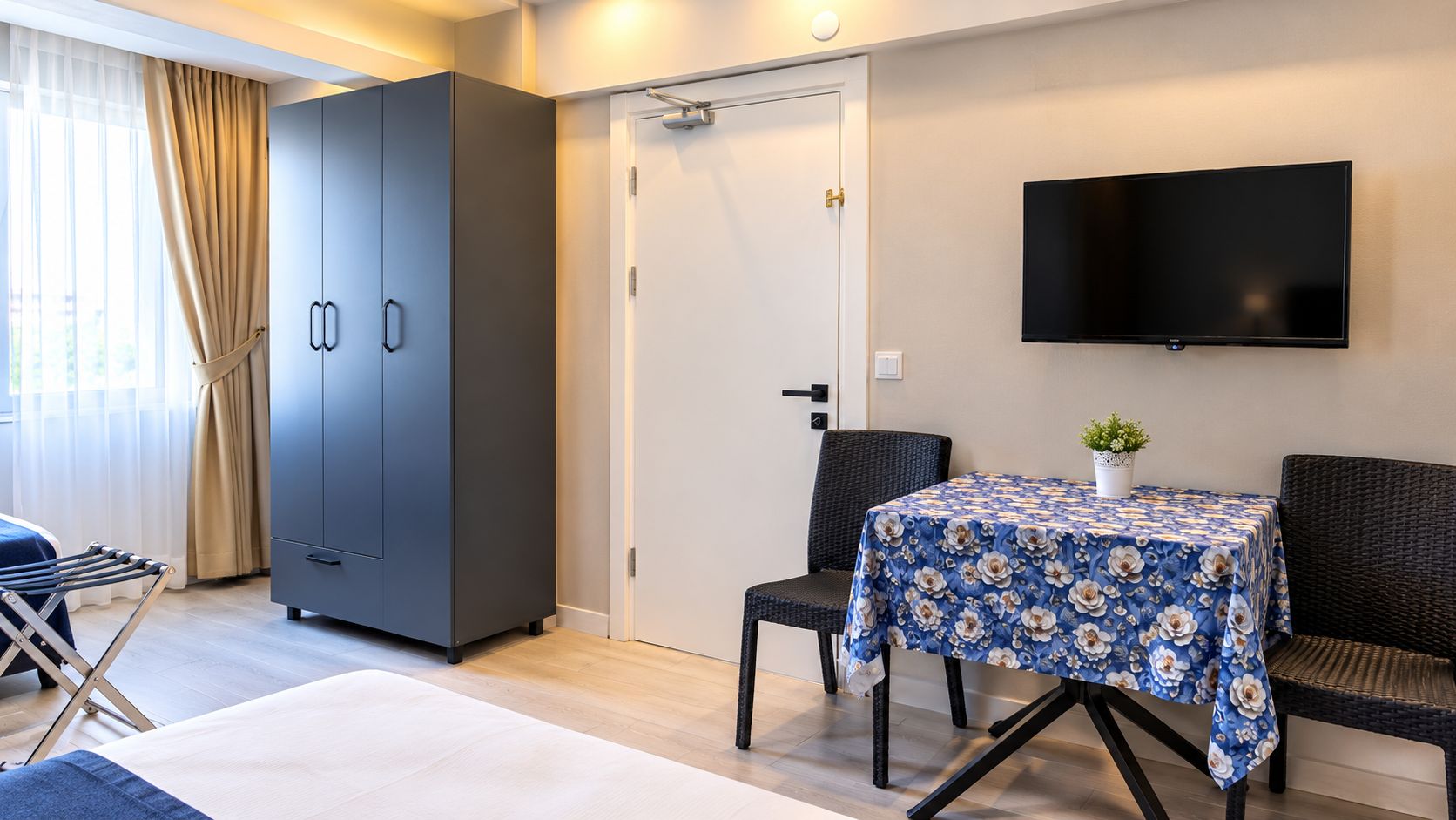 A hotel room with a bed, a wardrobe, a table with a floral tablecloth, a TV mounted on the wall, and a door with a towel rack beside it.