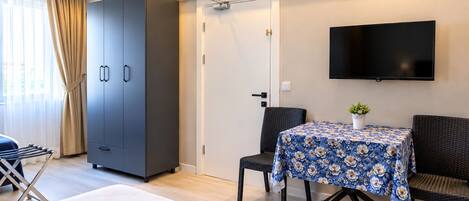 A hotel room with a bed, a wardrobe, a table with a floral tablecloth, a TV mounted on the wall, and a door with a towel rack beside it.