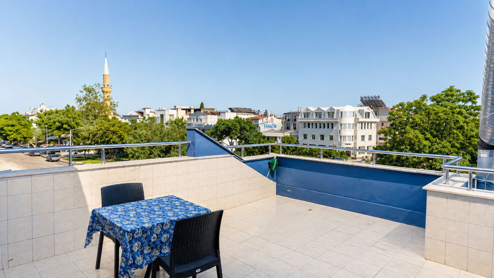 A rooftop terrace with a table and chairs, a blue railing, and a view of buildings and trees.