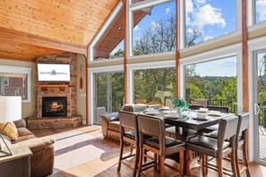 A spacious living area with a fireplace, a dining table set for four, and large windows offering a view of the outdoors.