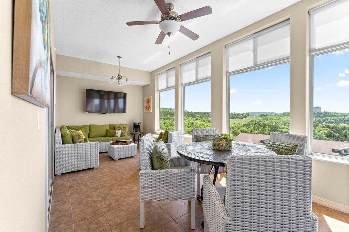 A spacious living area with a ceiling fan, a flat-screen TV, wicker furniture, and a large window offering a view of greenery.